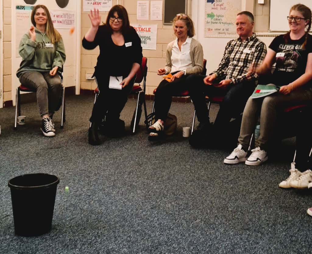 Donaldsons 1 A group of people are sat in a semicircle throwing scrunched up post it notes into a bin. They are laughing.