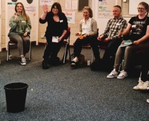 A group of people are sat in a semicircle throwing scrunched up post it notes into a bin. They are laughing.